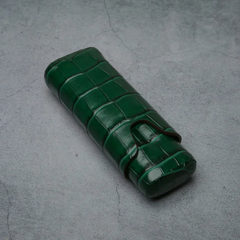 Emerald green croc-embossed leather cigar case with strap closure on textured gray surface
