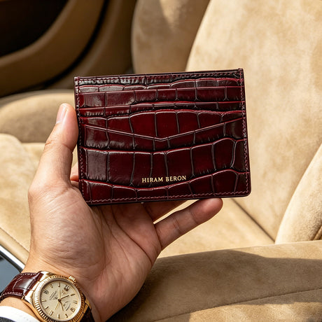 Hand holding a burgundy crocodile-patterned wallet with 'Hiram Beron' branding, against a beige background.