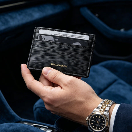 Hand holding a black EPI minimalist credit card holder with visible brand name, wearing a gold watch, against a dark background.