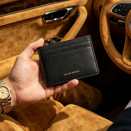 Hand holding a black saffiano leather cardholder with 'Hiram Beren' branding inside a car.