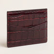 A glossy crocodile-embossed card holder wallet in deep burgundy, shown standing upright on a light beige background.