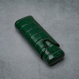 Emerald green croc-embossed leather cigar case with strap closure on textured gray surface