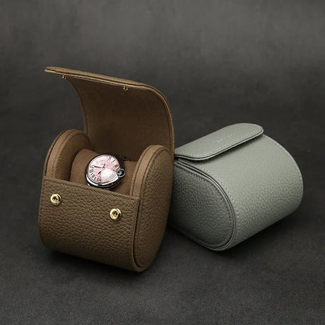 Etoupe pebble leather watch storage case with a watch inside, next to a gray watch roll case on a dark background